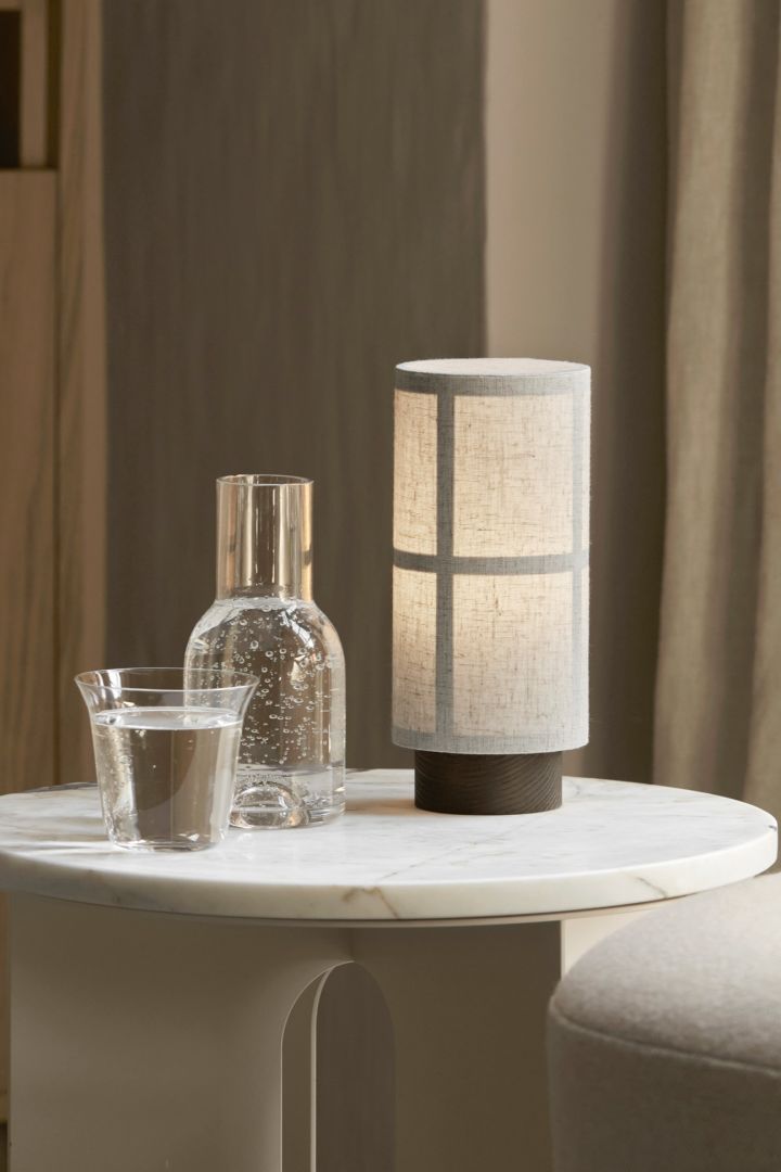 Hier sehen Sie die skandinavische Designlampe Hashira von Audo Copenhagen auf einem marmornen Beistelltisch stehen, daneben eine Karaffe und ein Glas mit Wasser.