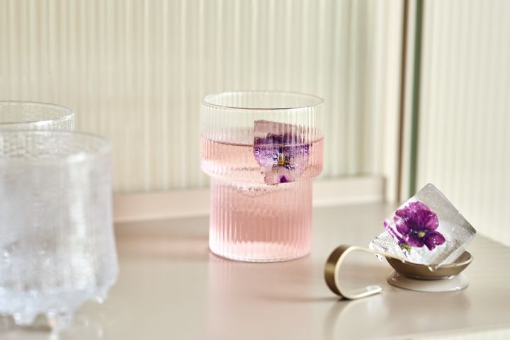 Ripple Trinkglas von Ferm Living mit einem rosa Getränk und einer lila Blume eingefroren in einem Eiswürfel.