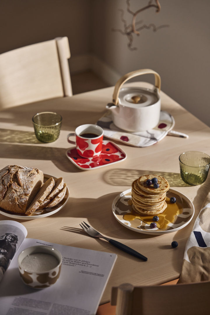 Hier sehen Sie einen Frühstückstisch mit den Marimekko Unikko-Tellern und der Teekanne sowie einem Stapel Pfannkuchen mit Sirup.