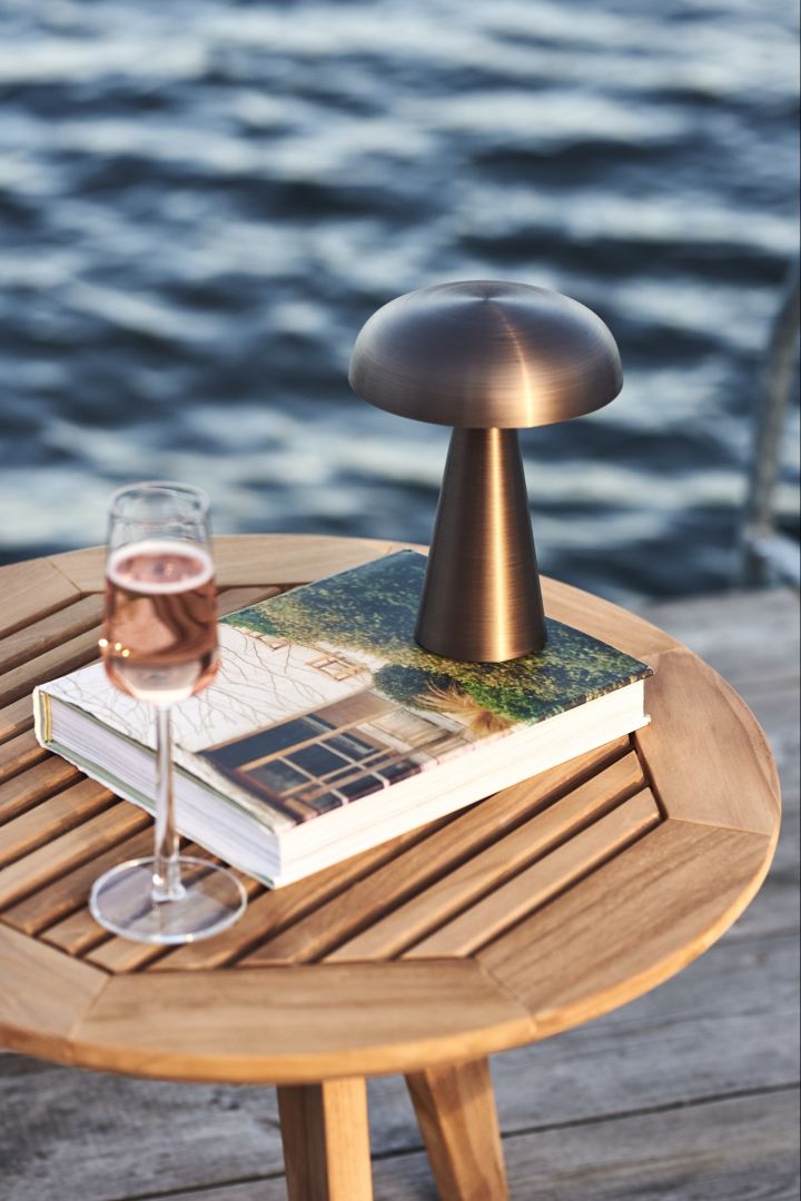 Hier sehen Sie eine kabellose Lampe, die Como von &Tradition, auf einem Holztisch, zusammen mit einem Glas Sekt, direkt am Wasser.