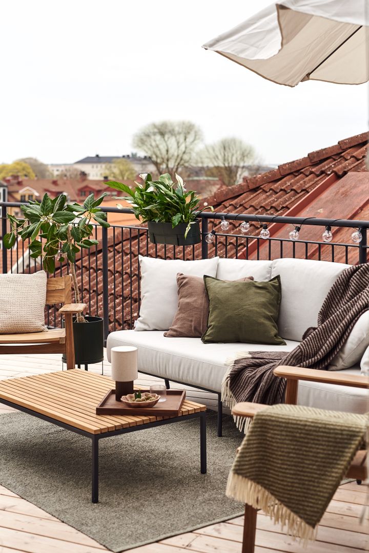 Das Bild zeigt einen Balkon, ausgestattet mit einem Sofa und Stühlen, Pflanzen, Kissen und Decken, die für eine gemütliche Atmosphäre sorgen.