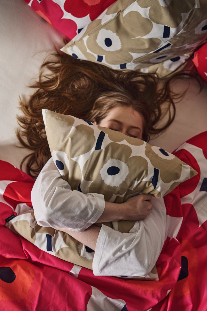 Eine Frau umarmt das Unikko-Kissen im Bett, umgeben von Unikko-Bettwäsche in Rot und Rosa.