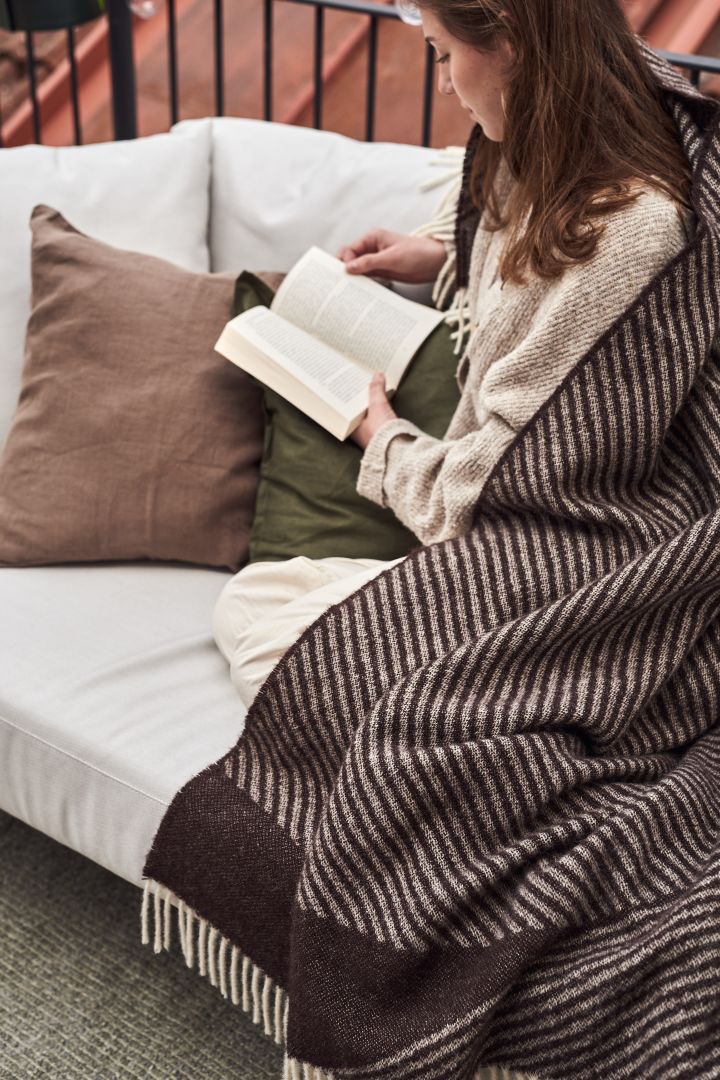 Hier sehen Sie eine Frau, die entspannt auf dem Outdoor-Sofa sitzt und ein Buch liest, eingewickelt in eine Stripes Wolldecke von NJRD.