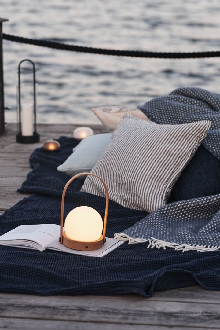 Die perfekten Begleiter für ein Picknick am Meer sind, wie hier zu sehen, ein paar Decken, Kissen und die kabellose Tischleuchte Carrie von Audo Copenhagen.